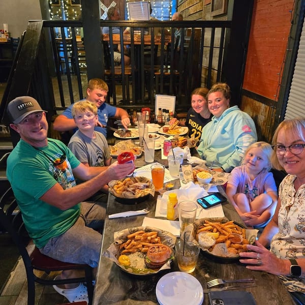 A family of seven smiles while dining at a restaurant with baskets of food and drinks on the table.