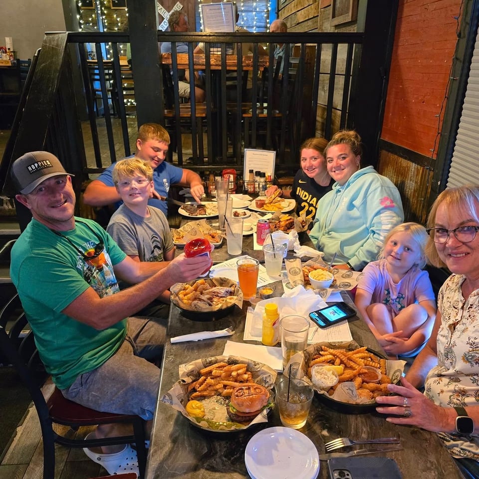 A family of seven smiles while dining at a restaurant with baskets of food and drinks on the table.