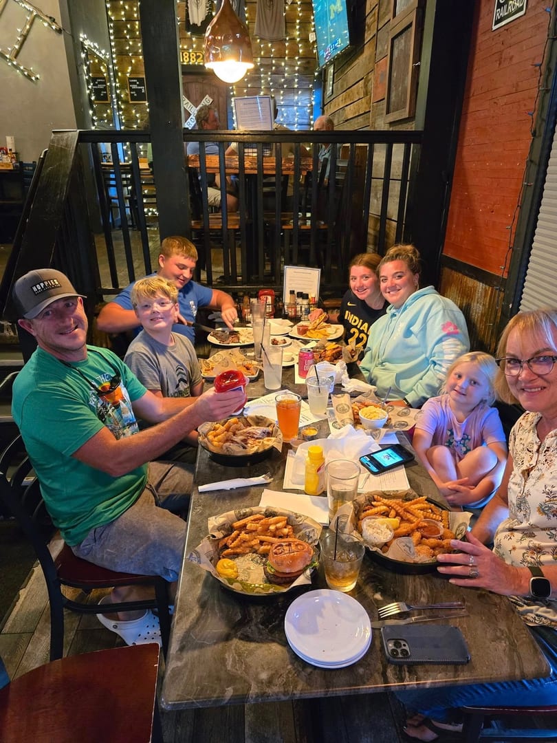 A family of seven smiles while dining at a restaurant with baskets of food and drinks on the table.