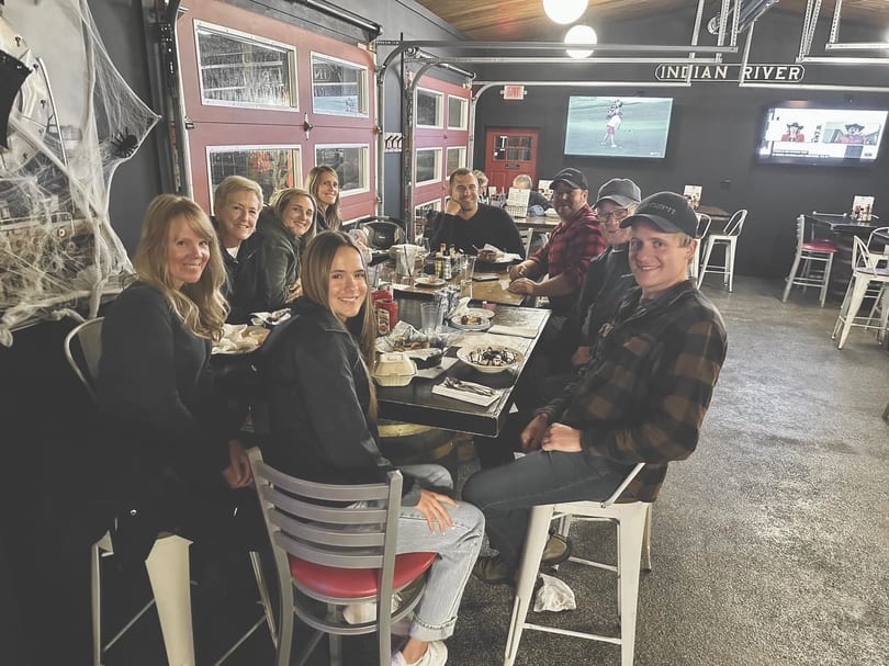 A group of people smiling and dining at a long table in a restaurant with Halloween decorations.