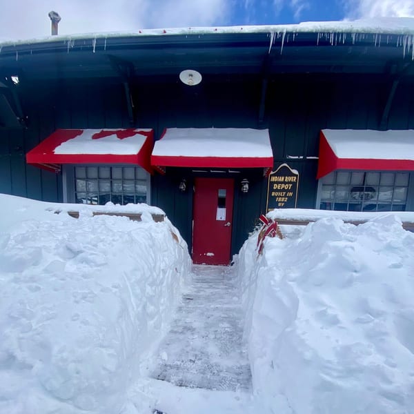 Snow-covered Indian River Depot building with red awnings and door, a cleared path leading to the entrance.