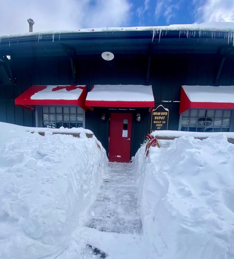 Snow-covered Indian River Depot building with red awnings and door, a cleared path leading to the entrance.