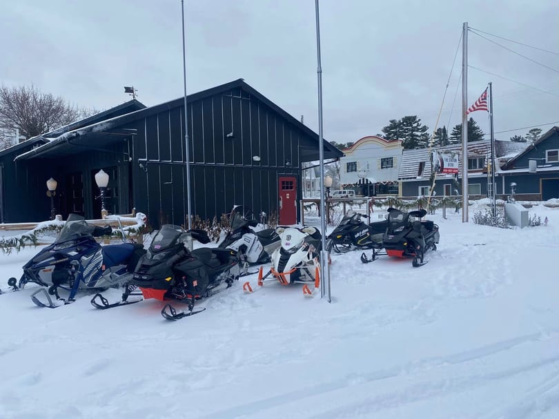 Snowmobiles parked in front of a dark building with a red door, surrounded by snow.