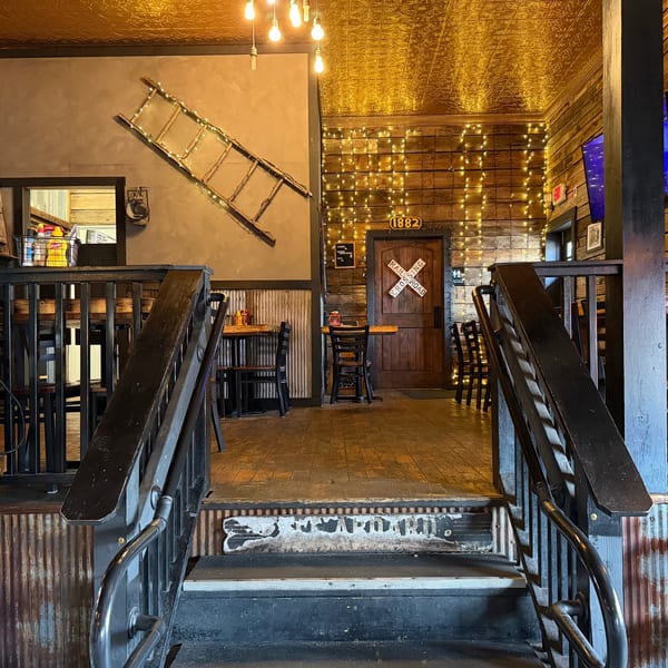 Rustic restaurant interior with wooden walls, tin ceiling, string lights, and a railroad crossing sign on a door.