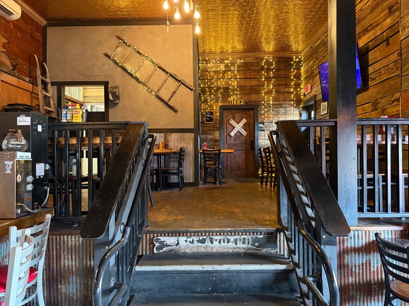Rustic restaurant interior with wooden walls, tin ceiling, string lights, and a railroad crossing sign on a door.