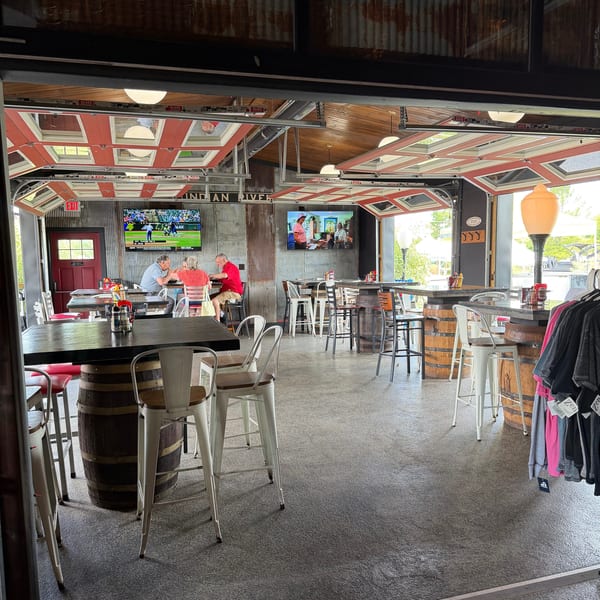 A casual restaurant interior with high-top tables, barrel bases, and TVs, with garage doors open.