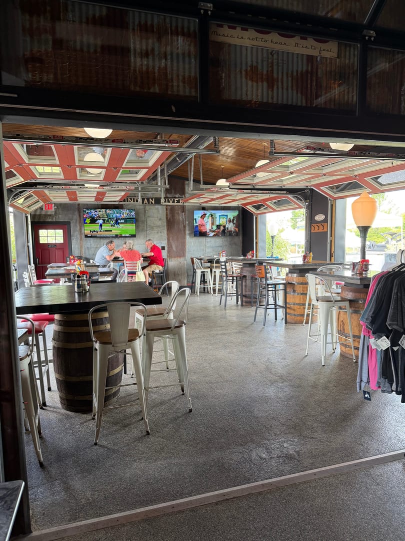 A casual restaurant interior with high-top tables, barrel bases, and TVs, with garage doors open.