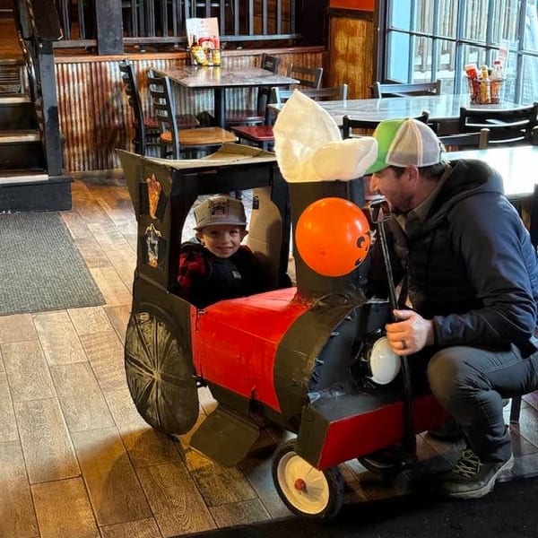 A young boy in a homemade train costume smiles at a man kneeling beside him in a restaurant.