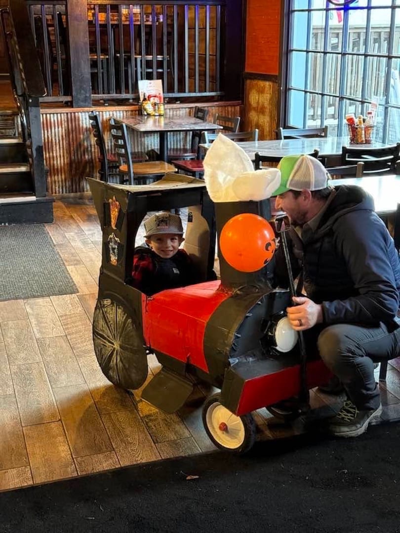 A young boy in a homemade train costume smiles at a man kneeling beside him in a restaurant.