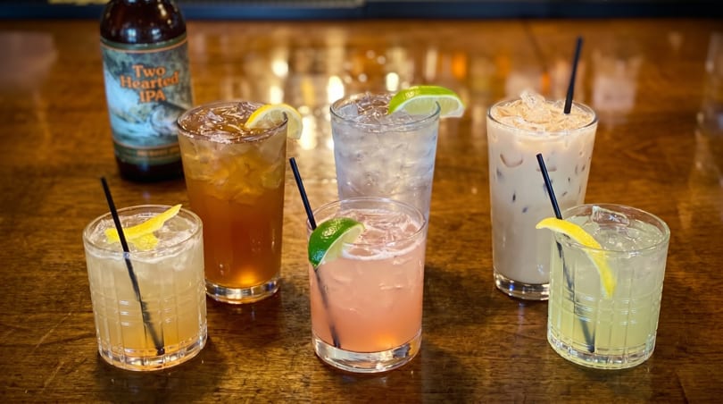 Six assorted cocktails and a bottle of Two Hearted IPA on a wooden bar.