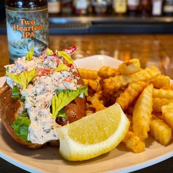 A lobster roll with fries, a lemon wedge, and a Two Hearted IPA beer bottle on a bar.