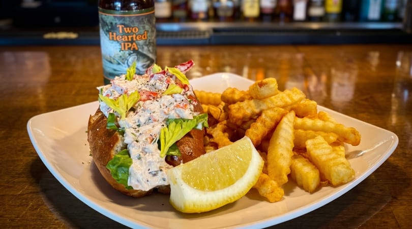 A lobster roll with fries, a lemon wedge, and a Two Hearted IPA beer bottle on a bar.