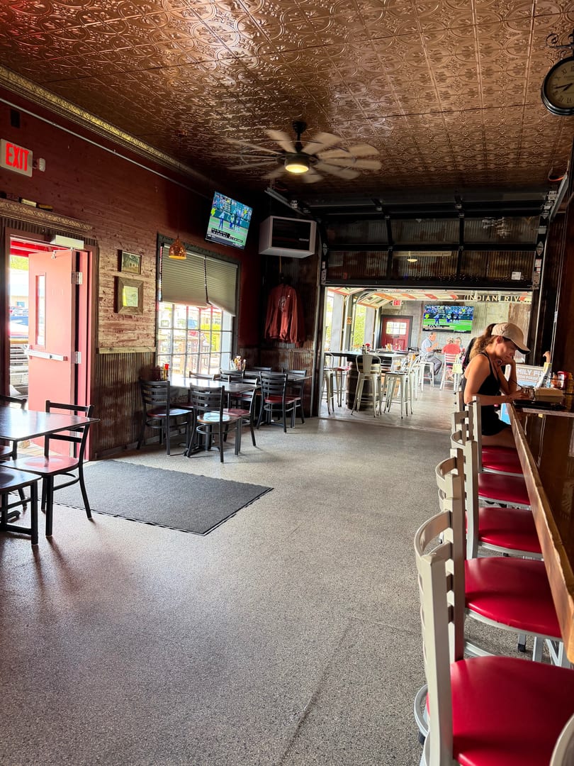 A woman sits at a bar in a rustic restaurant with a copper ceiling and open garage doors.