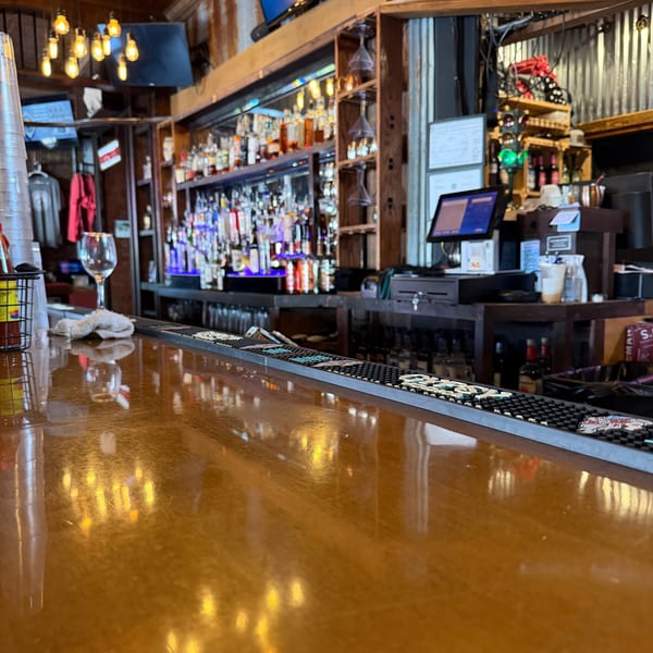 A close-up of a wooden bar top with reflections of Edison bulb lights, with a full bar in the background.