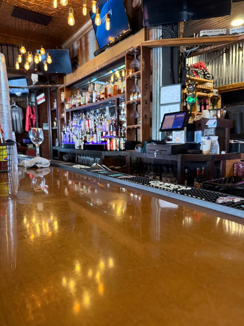 A close-up of a wooden bar top with reflections of Edison bulb lights, with a full bar in the background.